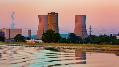 Kühltürme stoßen Dampf aus. Sie stehen in einer Industrieanlage mit Stromleitungen im Vordergrund. Der Himmel ist bei Sonnenuntergang in warmen Farben getaucht. Der Vordergrund zeigt Wasser mit sanften Wellen.