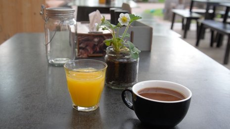 Ein Glas Orangensaft und eine Tasse Kaffee stehen auf einem Tisch in einem Café. Daneben sind ein Einmachglas mit einer Pflanze und Servietten zu sehen. Im Hintergrund Sitzgelegenheiten.