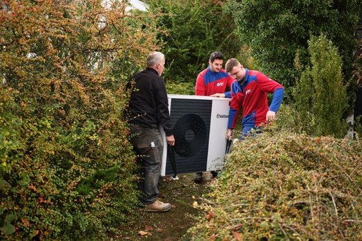 Drei Männer tragen eine Vaillant-Wärmepumpe durch einen dicht bewachsenen Garten. Sie tragen Arbeitskleidung, während sie das Gerät vorsichtig zwischen den Büschen manövrieren.