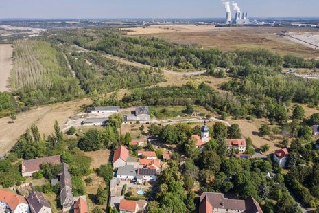 Luftaufnahme von einem Dorf bei Sonnenschein. Im Hintergrund ist ein kahles Kohlebaugebiet zu sehen.