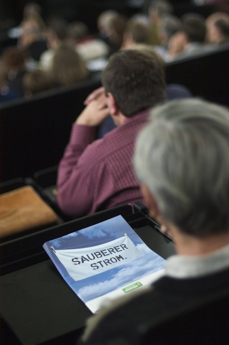 Person mit kurzen grauen Haaren sitzt in Vorlesungssaal. Vor ihr liegt ein Heft mit der Aufschrift "Sauberer Strom".
