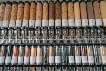 Spendersystem mit Körnern und Nudeln, geordnet in einem Geschäft. Die Beschriftungen zeigen Produktnamen und Preise. Die Umgebung ist ein moderner Laden für unverpackte Lebensmittel. Geeignet für nachhaltiges Einkaufen.