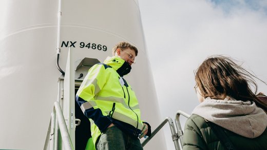 Mann in gelber Jacke vor dem Fuss eines Windrads im Gespräch mit einer weiteren Person.