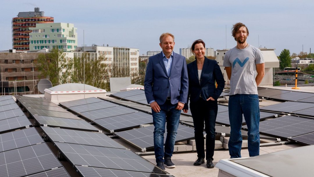 Drei Personen stehen auf einem Flachdach und lächeln in die Kamera. Um sie herum sind Solarpanelle zu sehen.