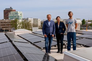 Drei Personen stehen auf einem Flachdach und lächeln in die Kamera. Um sie herum sind Solarpanelle zu sehen.