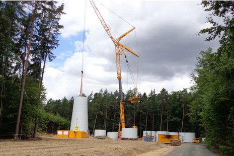Kran hebt einen Teil des Mastes der Windkraftanlage an. Im Hintergrund ein Wald.