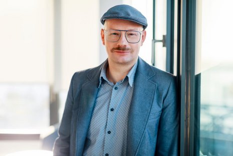 Mann mit graublauer Schiebermütze und gleichfarbigem Sakko steht an einem Fenster geleht und schaut freundlich in die Kamera. Er trägt einen kurzen Schnauzer.