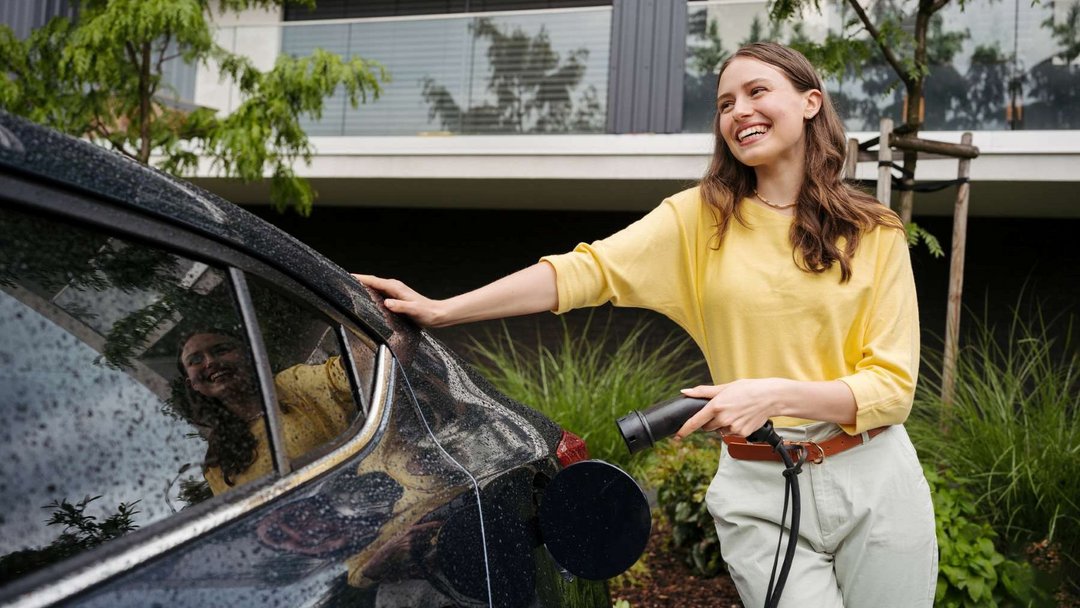 Eine Frau mit braunen langen Haaren und gelben Shirt steht vor einem begrünten Haus an einem schwarzen Auto und hält einen Ladestecker in der Hand.