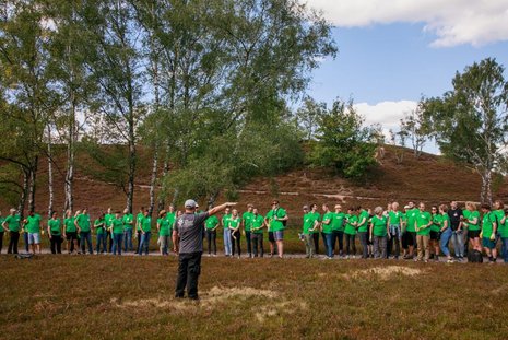 Eine Gruppe Menschen in grünen T-Shirts steht auf einem Heide-Feld im Halbkreis um eine Person in grauem Shirt herum. Diese zeigt mit der Hand in eine Richtung und scheint zu sprechen.