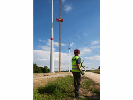 Ein Arbeiter mit Bauhelm und Warnweste sieht zu wie ein Baukran die Gondel eines Windrads nach oben befördert.
