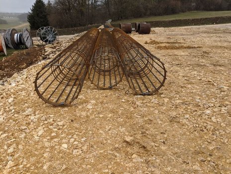 Drei große Metallgitterrohre liegen auf einem steinigen Untergrund. Im Hintergrund befinden sich mehrere Kabeltrommeln und eine grüne, bewaldete Landschaft.