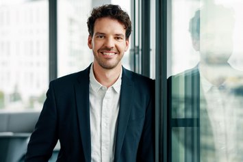 Ein braunhaariger Mann in weißem Hemd steht in einem modernen Büro an ein bodenlanges Fenster gelehnt und lächelt in die Kamera.