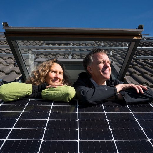 Ein Paar schaut aus dem Dachfenster heraus. Unter dem Fenster ist ein Solarpanel angebracht.