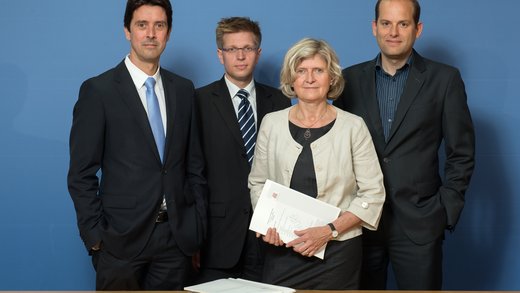 3 Männer und eine Frau, elegant gekleidet, schauen in die Kamera vor einem blauen Hintergrund.