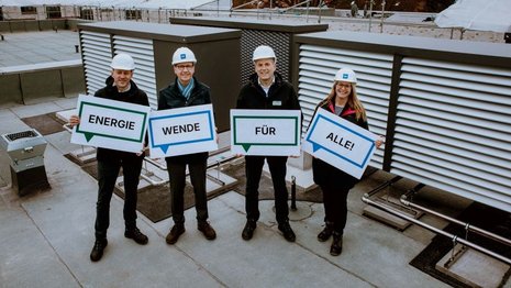 Auf einem Flachdach stehen vier Personen nebeneinander. Alle tragen weiße Schutzhelme und wetterfeste Kleidung. Jede Person hält ein Schild. Zusammen ergeben die Schilder den Satz "Energiewende für alle!". Im Hintergrund sind große Wärmepumpen zu sehen. Die Personen schauen in die Kamera und wirken freundlich und engagiert.