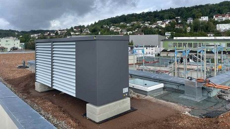 Wärmepumpenanlage auf dem Flachdach eines Gebäudes, montiert auf Betonsockeln. Im Hintergrund sind weitere Gebäudestrukturen, Leitungsführungen und ein städtische Hanglage mit Wohnhäusern zu sehen. Über der Szene hängen dunkle Wolken.