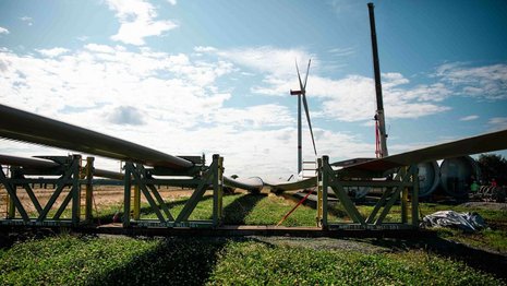 Windturbinenflügel liegen auf Gestellen, während ein Kran im Hintergrund eine Turbine errichtet. Die Szene spielt auf einem grünen Feld unter blauem Himmel.