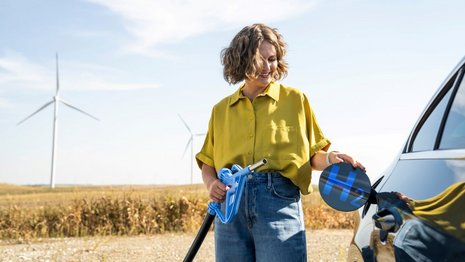 Eine junge Frau mit kurzen blonden Haaren steht an einem Weizenfeld und tankt ein Auto. Auf der Tankklappe steht "H2". Im Hintergrund sind Windräder zu sehen.