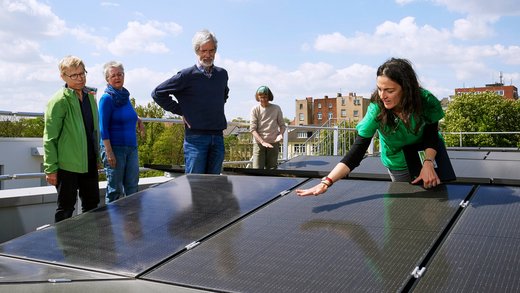 Eine Gruppe Menschen steht um PV-Module auf einem Flachdach. Eine Frau mit dunkeln langen Haaren streicht über eines rüber.
