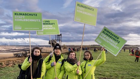Vier Personen stehen auf einer Wiese und halten Schilder mit der Aufschrift „Klimaschutz statt Kohleschmutz“ und „Wirksamer Klimaschutz für globale Gerechtigkeit“. Im Hintergrund ist ein Tagebau mit Baggern sichtbar.