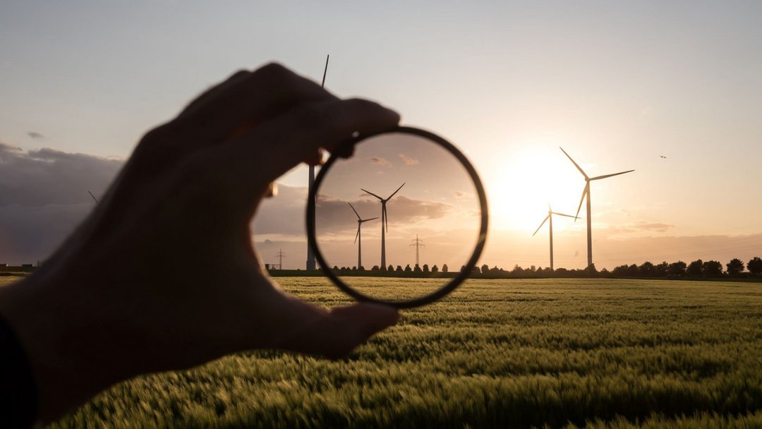 Windkraftanlagen auf einem Feld bei Sonnenuntergang. Eine im Schatten liegende Hand hält ein rundes lupenähnliches Glas vor die Windräder.