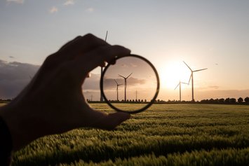 Windkraftanlagen auf einem Feld bei Sonnenuntergang. Eine im Schatten liegende Hand hält ein rundes lupenähnliches Glas vor die Windräder.