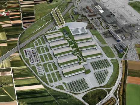 Eine Luftaufnahme eines Messegeländes.Die Dächer der Messehallen sind teilweise gegrünt. Das Gelände ist von Grünanlagen und Feldern umschlossen. An das Gelände grenzt ein Flughafen.