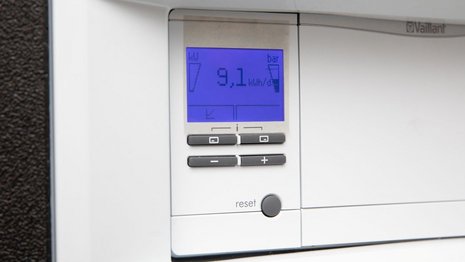 Ein digitales Display eines Heizsystems zeigt "9,1 kWh/d" an. Darunter befinden sich Steuerungstasten in einem weißen Gehäuse. Umgebung: modern und funktional. Text: "reset".