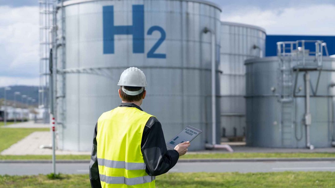 Ein Mann von hinten in gelber Sicherheitsweste hält ein Tablet in der Hand und sieht in Richtung einer Art Tank oder Industriecontainter auf dem H2 steht.