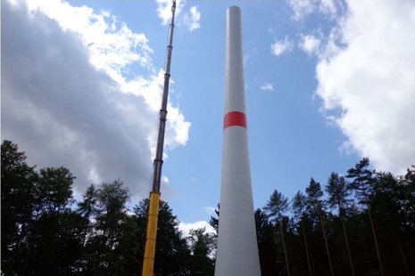 Blick von unten auf einen Kran, der einen Windkraftanlagenmast montiert. Im Hintergrund ein Wald.