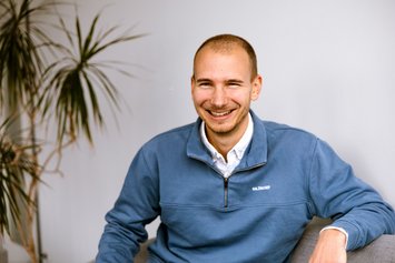 Ein Mann mit sehr kurzen Haaren und leichtem Bart sitzt auf einem grauen Sofa. Im Hintergrund ist eine Zimmerpalme zu sehen. Er trägt einen hellblauen Troyerpullover mit weißem Hemd darunter.