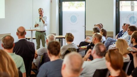 Ein man steht mit einem Mikrofon vor einer kleinen Gruppe und hält einen Vortrag. 