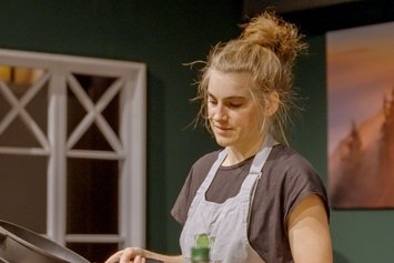 Eine Frau mit blonden, hochgebundenen Haaren trägt eine Schürze und hält eine Pfanne in der Hand