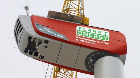 Aufnahme von unten der Montage des Rotors auf dem Mast einer Windkraftanlage mit einem gelben Kran.