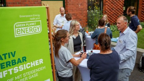Eine Gruppe verschiedener Menschen steht um einen weißen Stehtisch herum und unterhält sich. Im Vordergrund ist ein Green-Planet-Energy Banner zu sehen. 