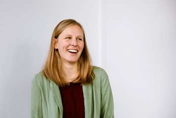 Eine junge Frau mit blonden langen Haaren lacht an der Kamera vorbei. Sie trägt eine hellgrüne Strickjacke und ein schwarzes Oberteil darunter.