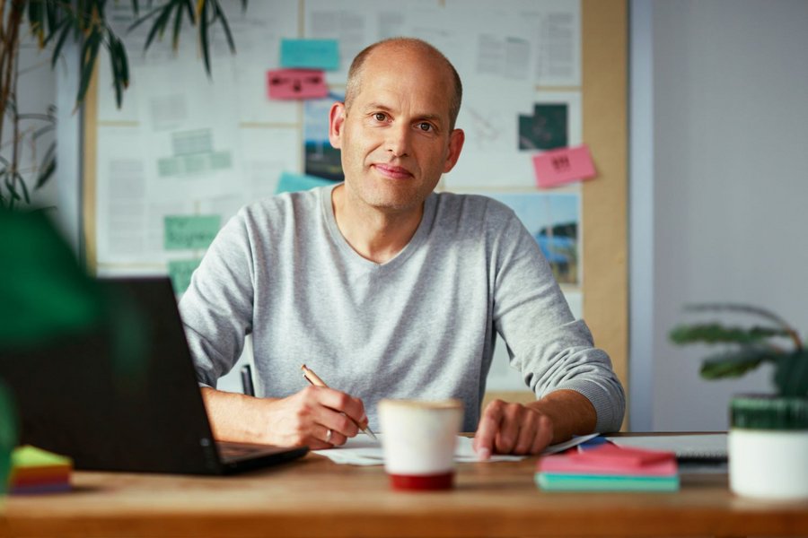 Ein Mann mit Halbglatze sitzt an einem Holztisch. Er trägt ein hellgraues Sweatshirt und blick freundlich in die Kamera. Im Hintergrund ist eine Pinnwand mit Postits und Zetteln zu sehen.