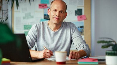 Ein Mann mit Halbglatze sitzt an einem Holztisch. Er trägt ein hellgraues Sweatshirt und blick freundlich in die Kamera. Im Hintergrund ist eine Pinnwand mit Postits und Zetteln zu sehen.