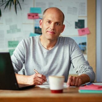 Ein Mann mit Halbglatze sitzt an einem Holztisch. Er trägt ein hellgraues Sweatshirt und blick freundlich in die Kamera. Im Hintergrund ist eine Pinnwand mit Postits und Zetteln zu sehen.