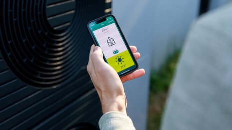 Kamerapersüektive über die Schulter einer Person in hellgrauem Pullover. Sie hält ein Smartphone in der Hand auf dem eine App eine Temperatur und Haus-Icon anzeigt. Im Hintergrund steht eine Wärmepumpe.