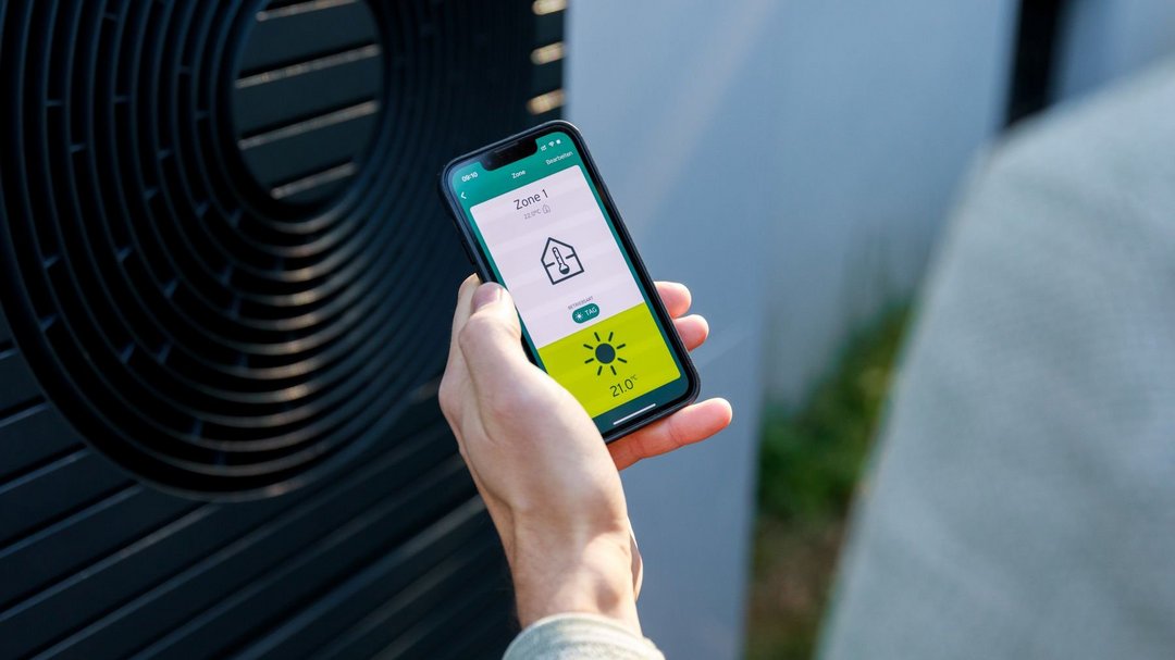 Kamerapersüektive über die Schulter einer Person in hellgrauem Pullover. Sie hält ein Smartphone in der Hand auf dem eine App eine Temperatur und Haus-Icon anzeigt. Im Hintergrund steht eine Wärmepumpe.