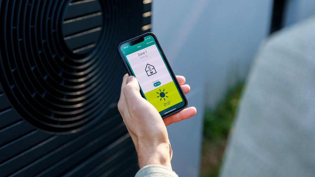 Kamerapersüektive über die Schulter einer Person in hellgrauem Pullover. Sie hält ein Smartphone in der Hand auf dem eine App eine Temperatur und Haus-Icon anzeigt. Im Hintergrund steht eine Wärmepumpe.