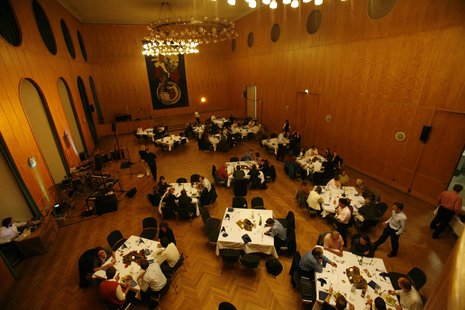 Großer Saal mit Kronleuchter und hölzerner Verkleidung von oben aufgenommen. Im Saal sind quadratische Esstische aufgebaut, an denen Menschen Essen zu sich nehmen.