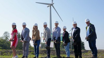 Eine Gruppe von acht Personen mit Helmen hält Spaten bei einem symbolischen Spatenstich vor einem Windrad auf einem offenen Feld unter klarem Himmel.
