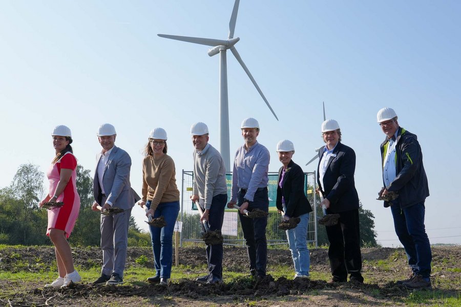 Eine Gruppe von acht Personen mit Helmen hält Spaten bei einem symbolischen Spatenstich vor einem Windrad auf einem offenen Feld unter klarem Himmel.