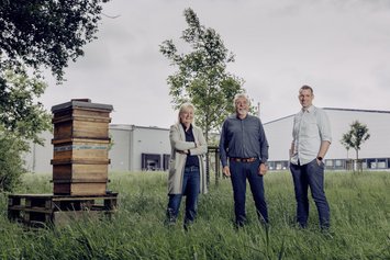 Zwei Männer und eine Frau stehen auf einer Wiese, neben ihnen stehen mehrere Holzkisten auf einer Holzpalette; im Hintergrund ein Firmengeäude.