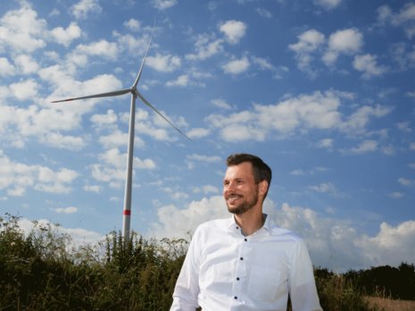 Ein lächelnder Mann in weißem Hemd steht vor einem Windrad.