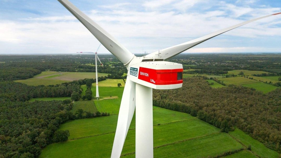 Nahe Drohnenaufnahme einer Windkraftanlage von hinten. An der linken Seite des Windradkopfes ist ein dunkelgrünes Logo mit der Aufschrift "Green Planet Energy" zu sehen.