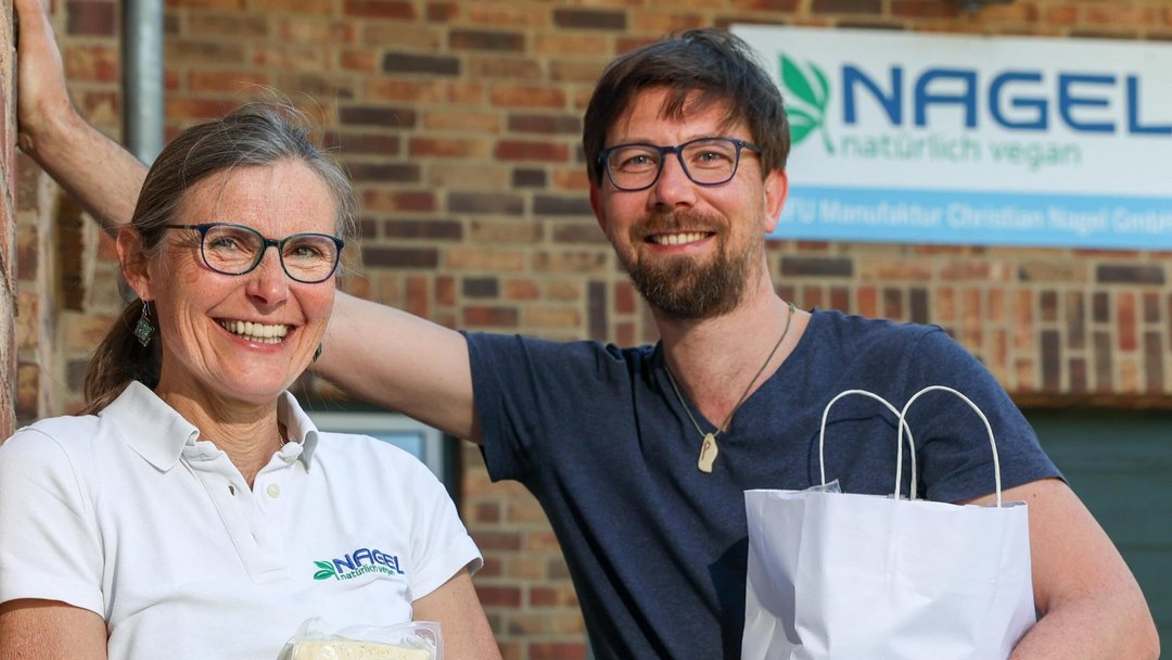 Eine Frau in weißem T-Shirt und ein Mann in blauem T-Shirt, beide mit Brille stehen vor einer Backsteinmauer mit dem Schild "NAGEL natürlich vegan". In der Hand halten Sie eine Tüte und eine Packung Tofu.