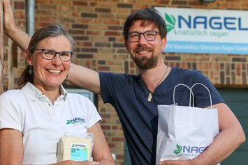 Eine Frau in weißem T-Shirt und ein Mann in blauem T-Shirt, beide mit Brille stehen vor einer Backsteinmauer mit dem Schild "NAGEL natürlich vegan". In der Hand halten Sie eine Tüte und eine Packung Tofu.